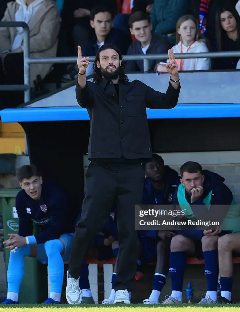 A man in a black jacket and black trousers stands on a sports field sideline gesturing with both hands, with teammates sitting on a bench behind him and spectators in the stadium stands.