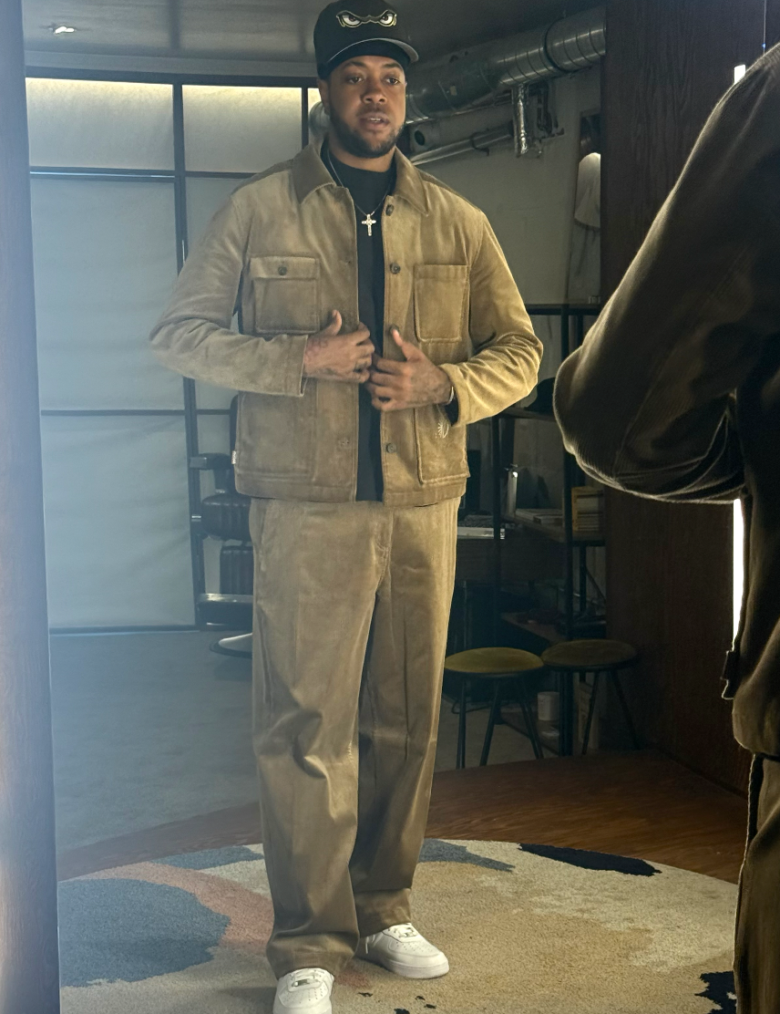 A man wearing a beige corduroy jacket and matching beige corduroy pants, styled with a black t-shirt, white sneakers, and a black cap, standing in a room with a mirror.