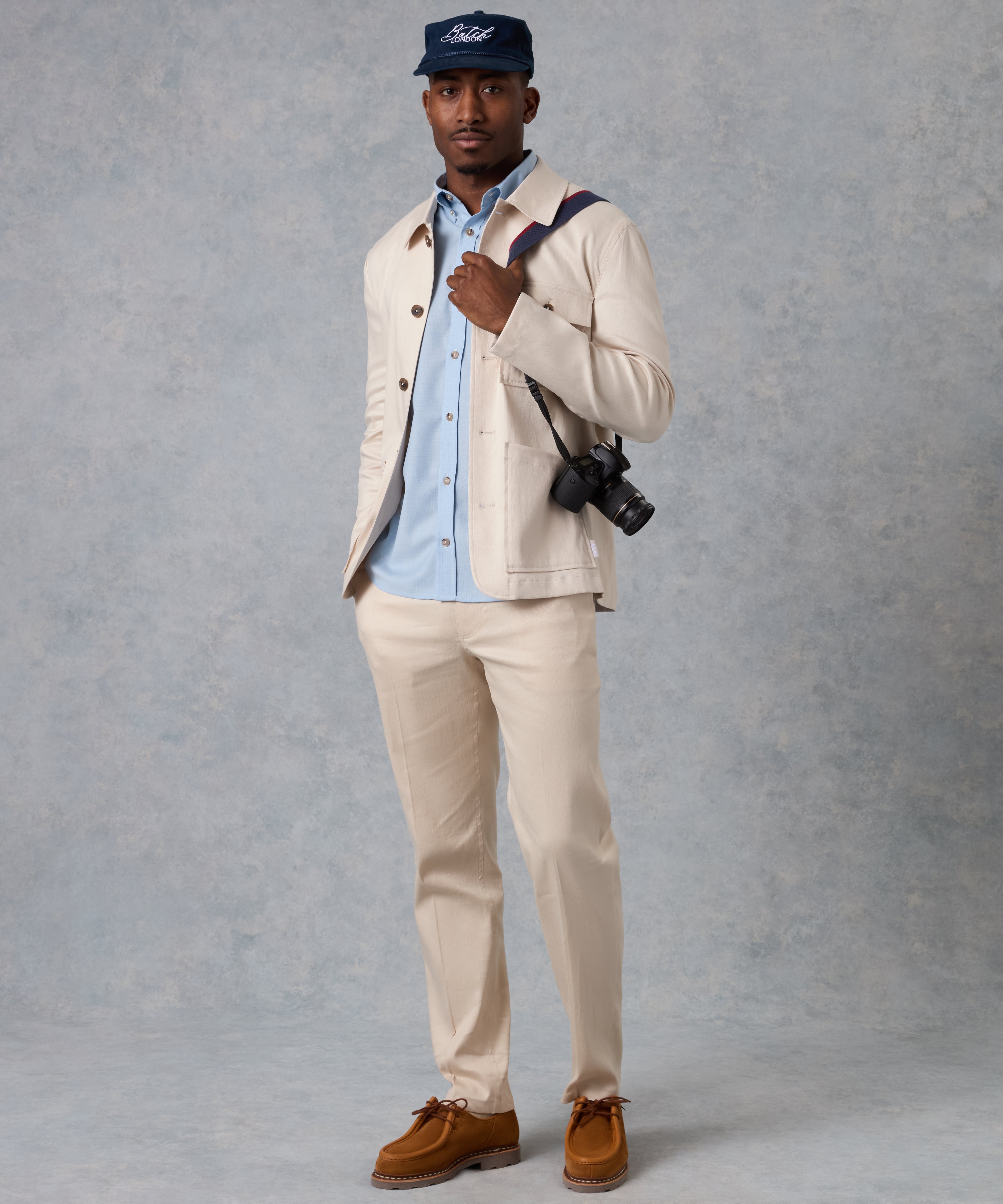 A man modeling a light beige two-piece suit with a light blue button-down shirt, a navy blue Ford cap, and brown suede shoes, standing against a textured gray studio backdrop.