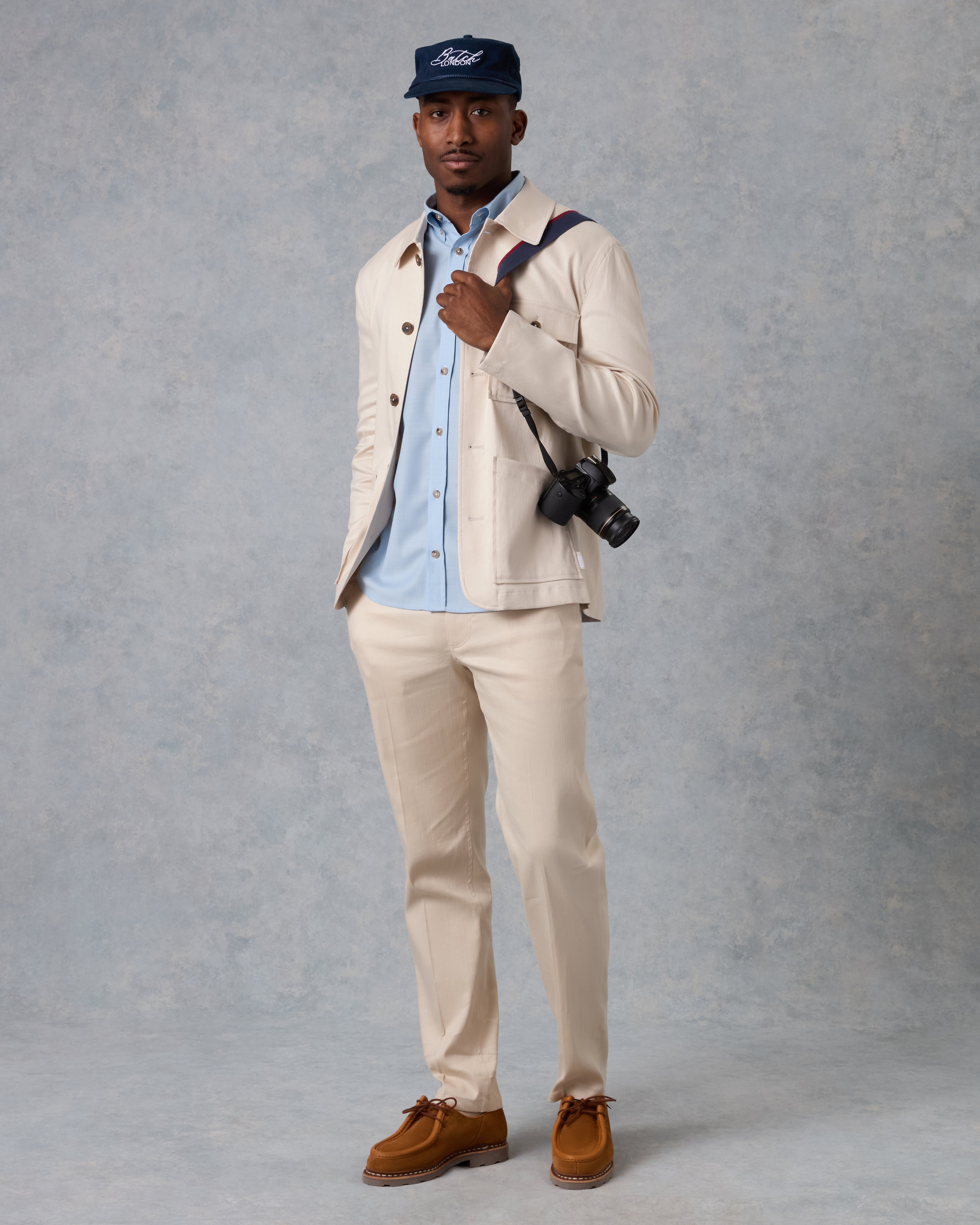 A man modeling a light beige two-piece suit with a light blue button-down shirt, a navy blue Ford cap, and brown suede shoes, standing against a textured gray studio backdrop.