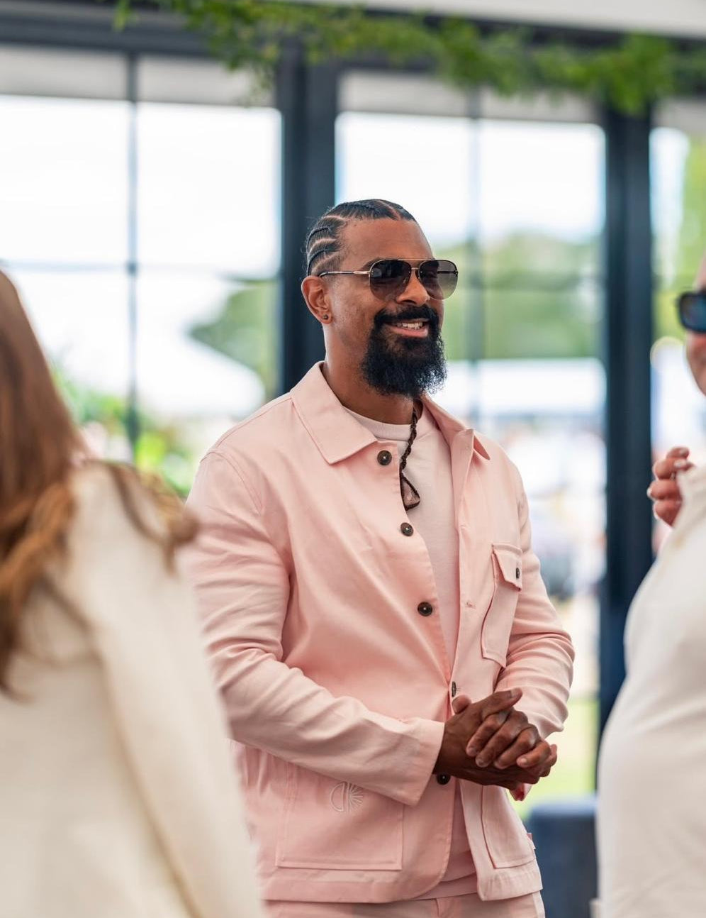 A man wearing a light pink two-piece suit and sunglasses, standing in a bright, modern indoor setting with large windows and greenery.