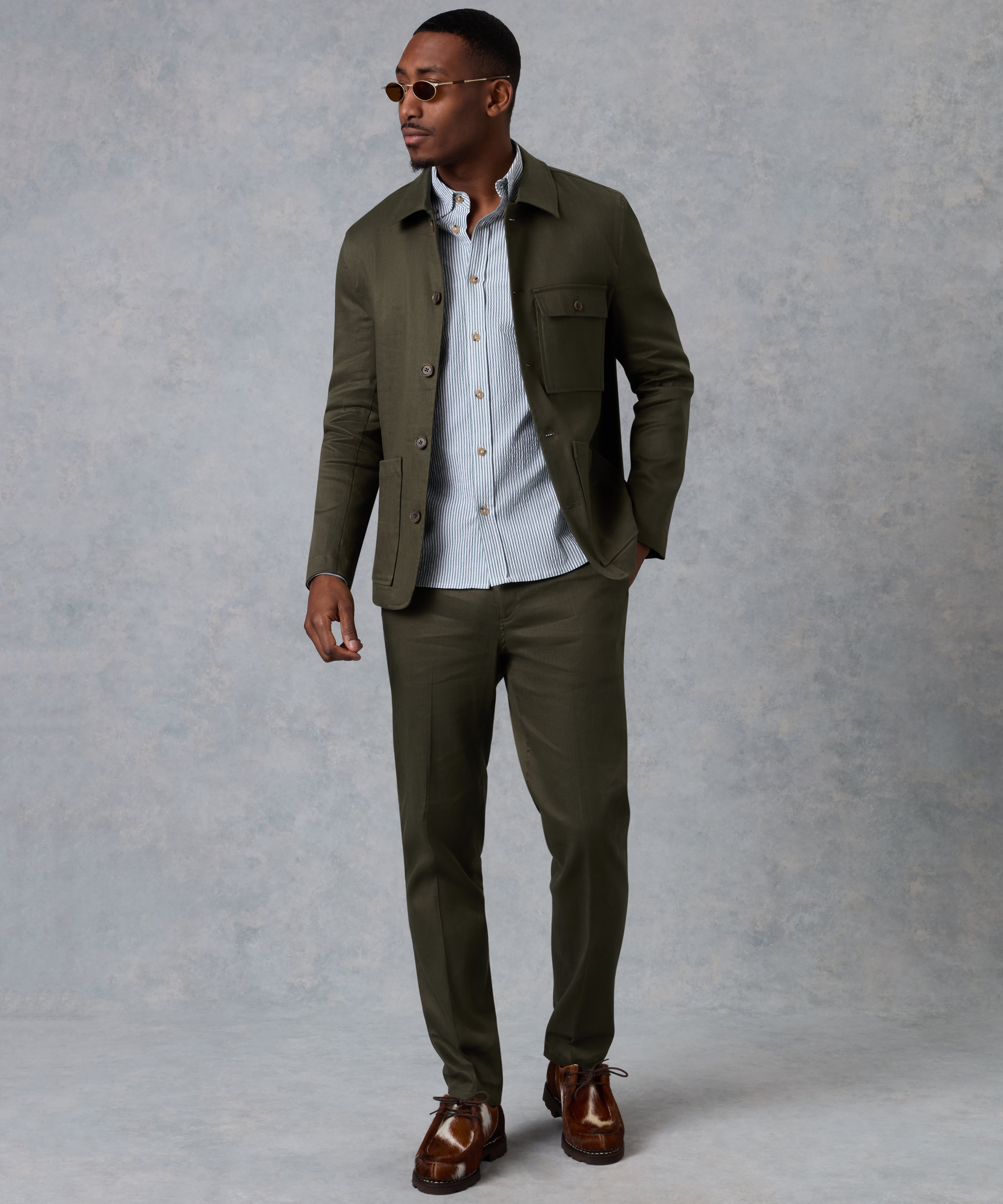 Man wearing an olive green jacket and pants with a light blue shirt against a gray background