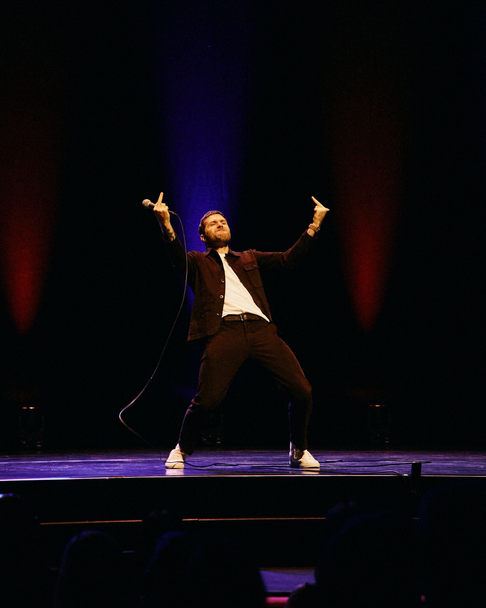 A man performing on a stage with arms raised, wearing a dark jacket and trousers over a white shirt, under dramatic red and blue stage lights.