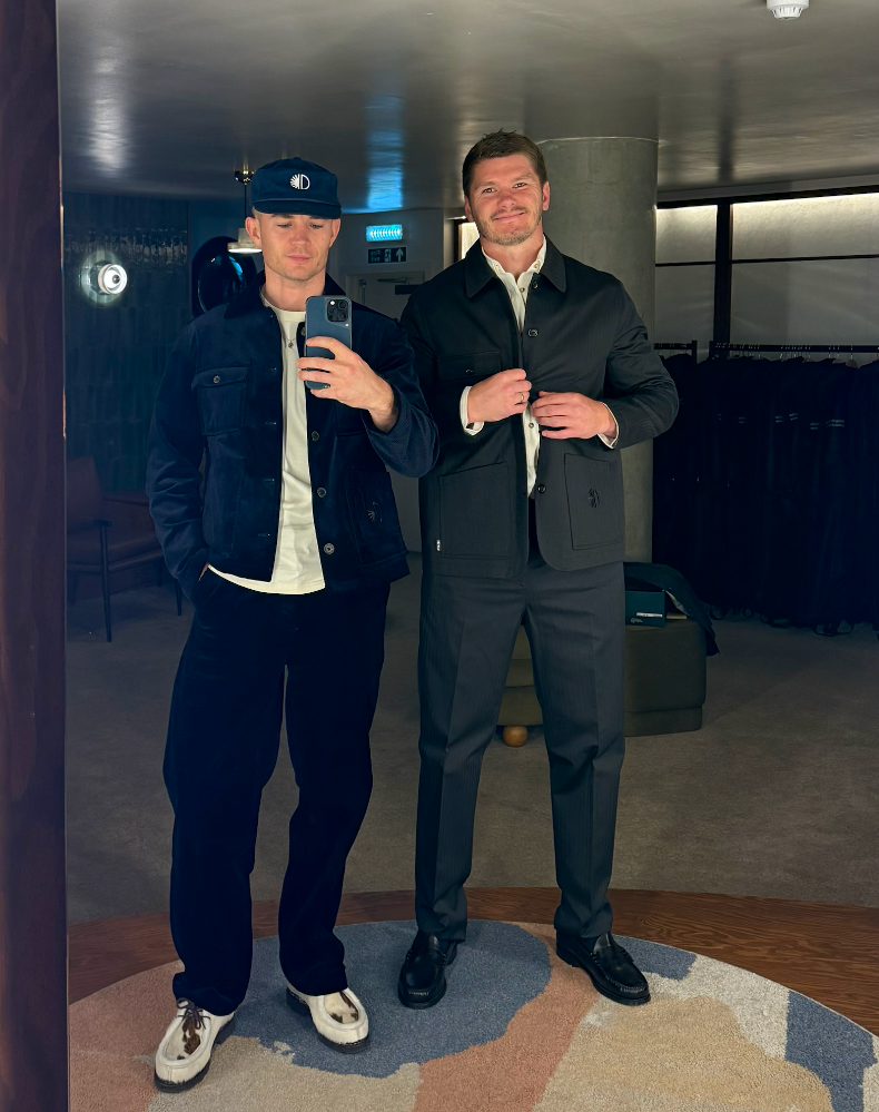 Two men posing for a mirror selfie in an indoor setting. The man on the left wears a dark blue corduroy jacket, black trousers, a white t-shirt, and a black cap.