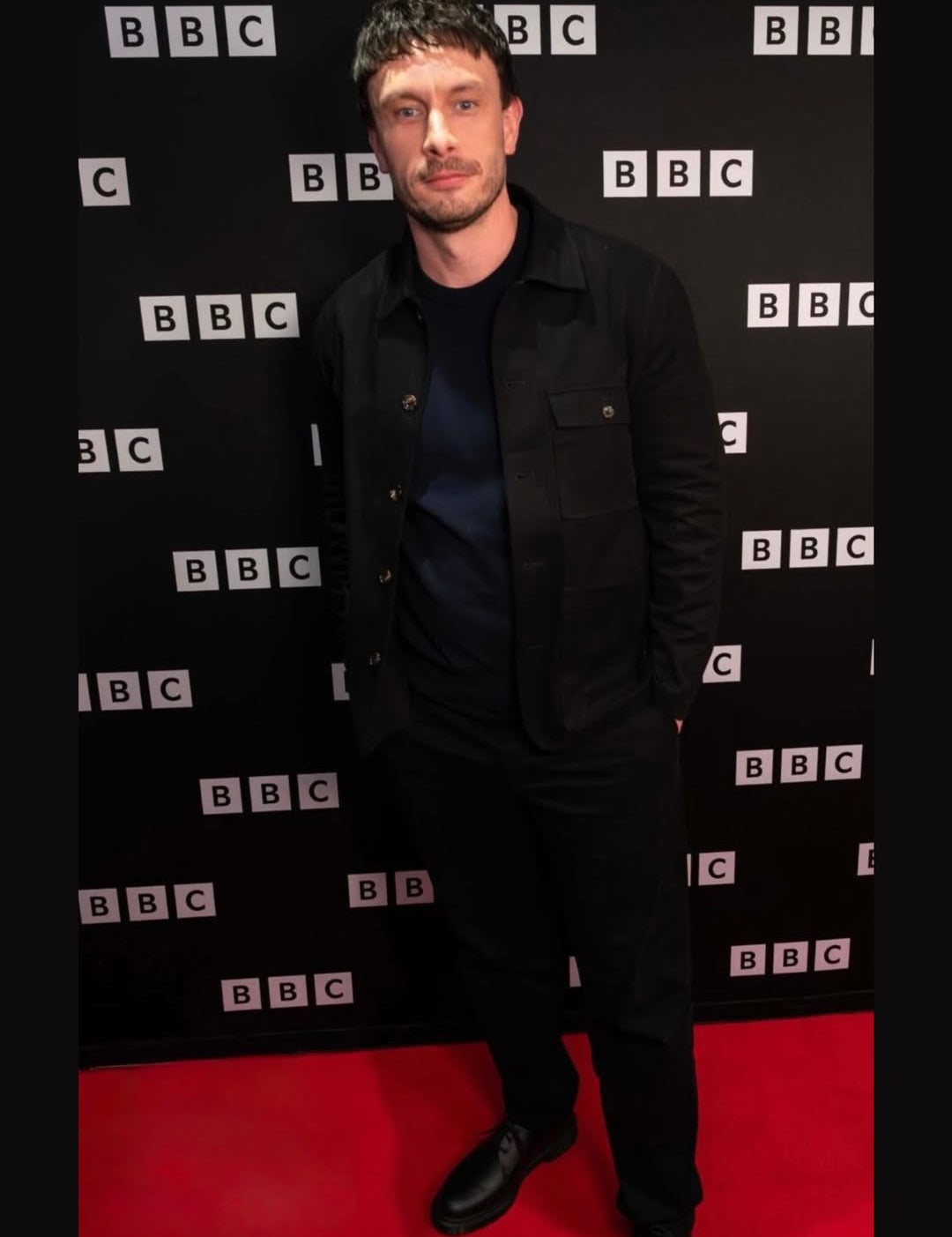 A man wearing a black jacket, dark blue t-shirt, and black trousers stands on a red carpet in front of a black BBC backdrop.
