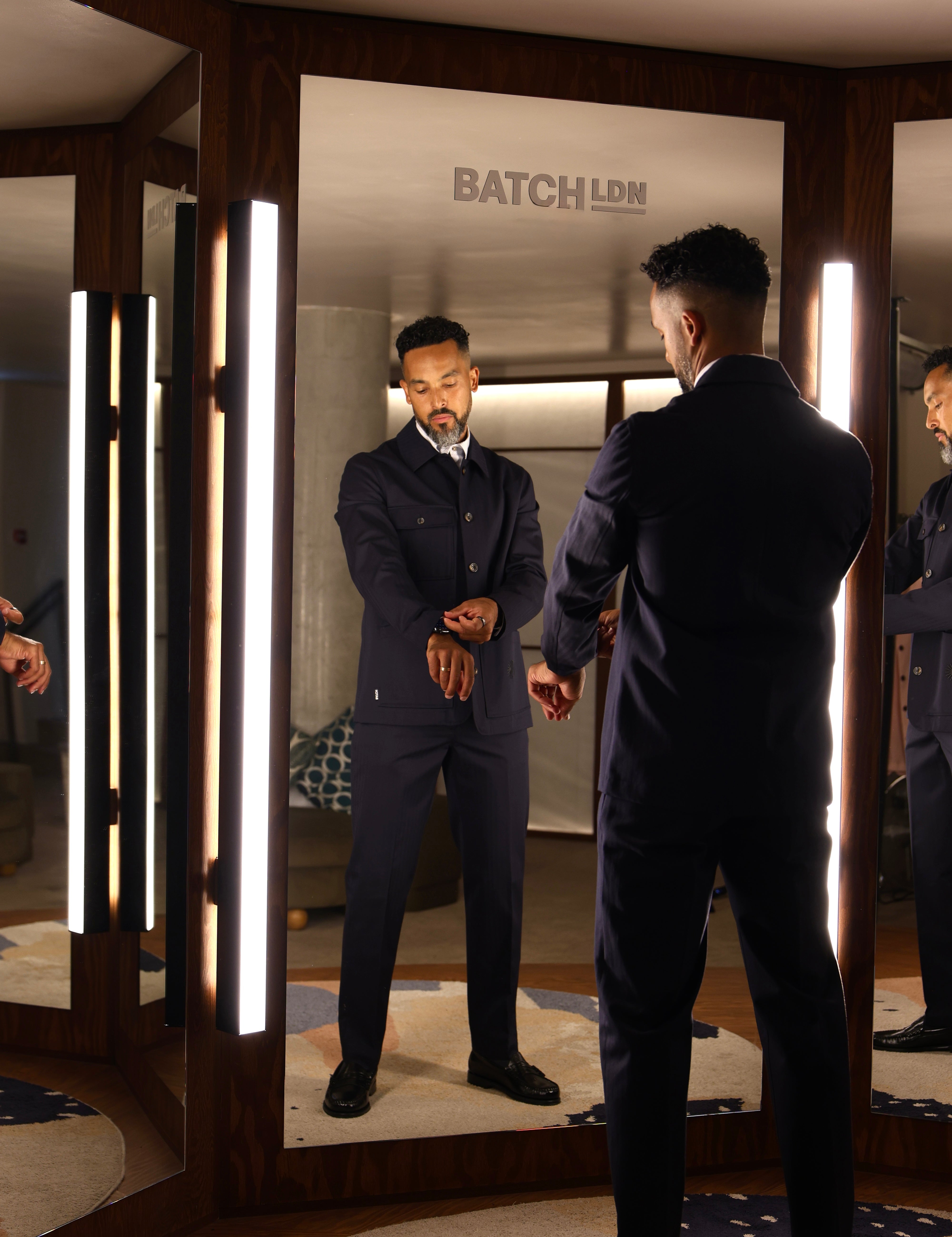 A man in a dark navy suit stands in front of a mirror in a modern fitting room, looking at his reflection. The room features wood paneling, vertical light strips, and a visible 'BATCH LDN' logo on the back of the mirror frame.